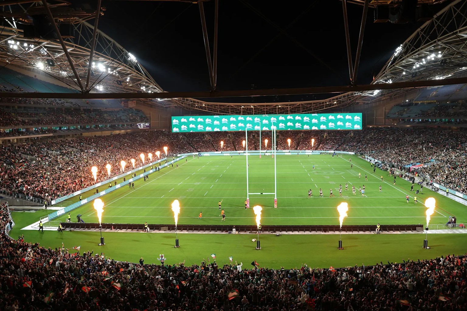A packed Accor Stadium at night, flame jets firing along the sideline as a match gets under way under floodlights