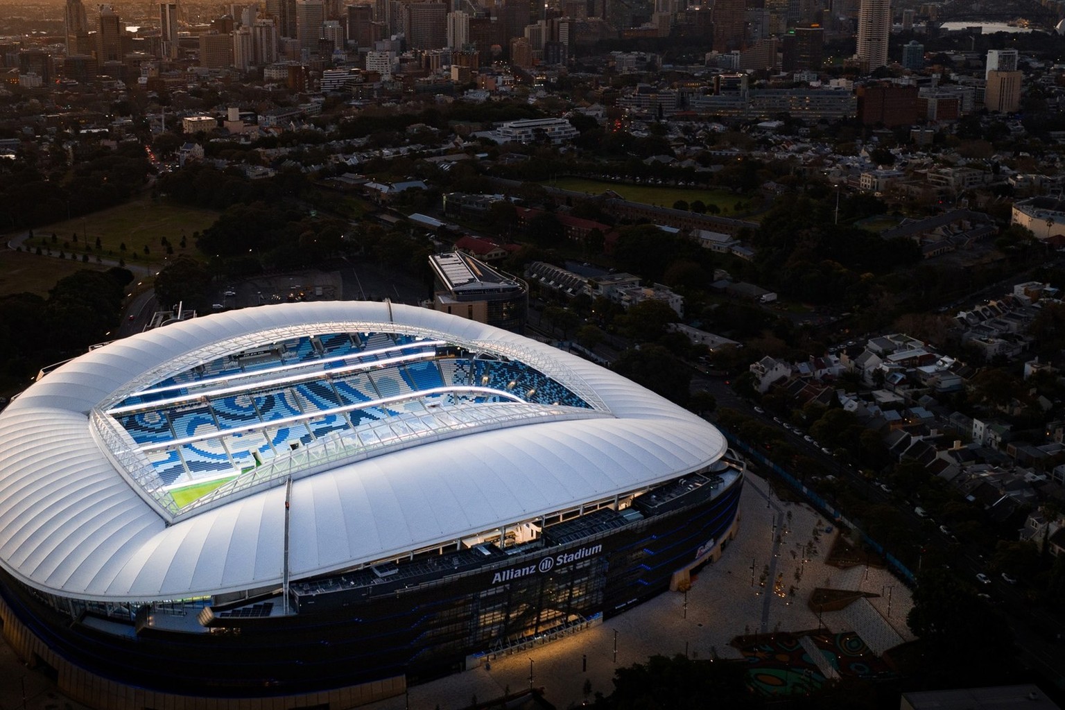 Allianz Stadium from the air in daylight, its white louvred roof ringing the pitch with the Sydney skyline behind it