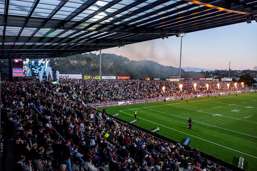 Brookvale Oval, 4 Pines Park, with a full grandstand and flame jets on the field at dusk, bushland on the hill beyond