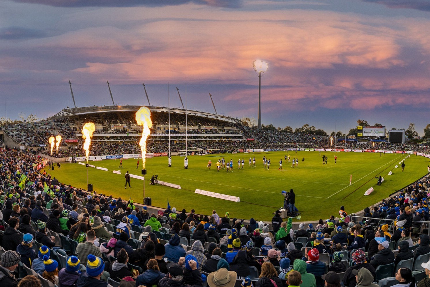 GIO Stadium Canberra at dusk under a pink sky, the crowd close to the field and flames firing as the teams take the field