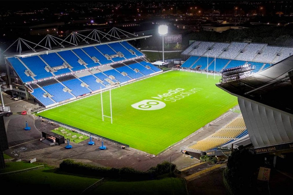 Go Media Stadium at night from above, the floodlit green pitch bright against dark blue seating