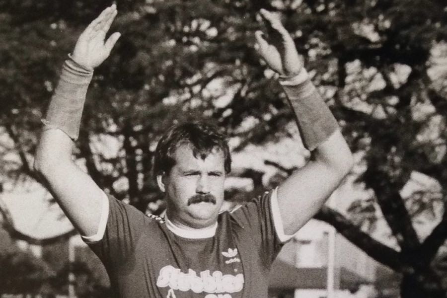 A moustachioed rugby league player in a Stubbies jersey raises both arms above his head on a suburban ground