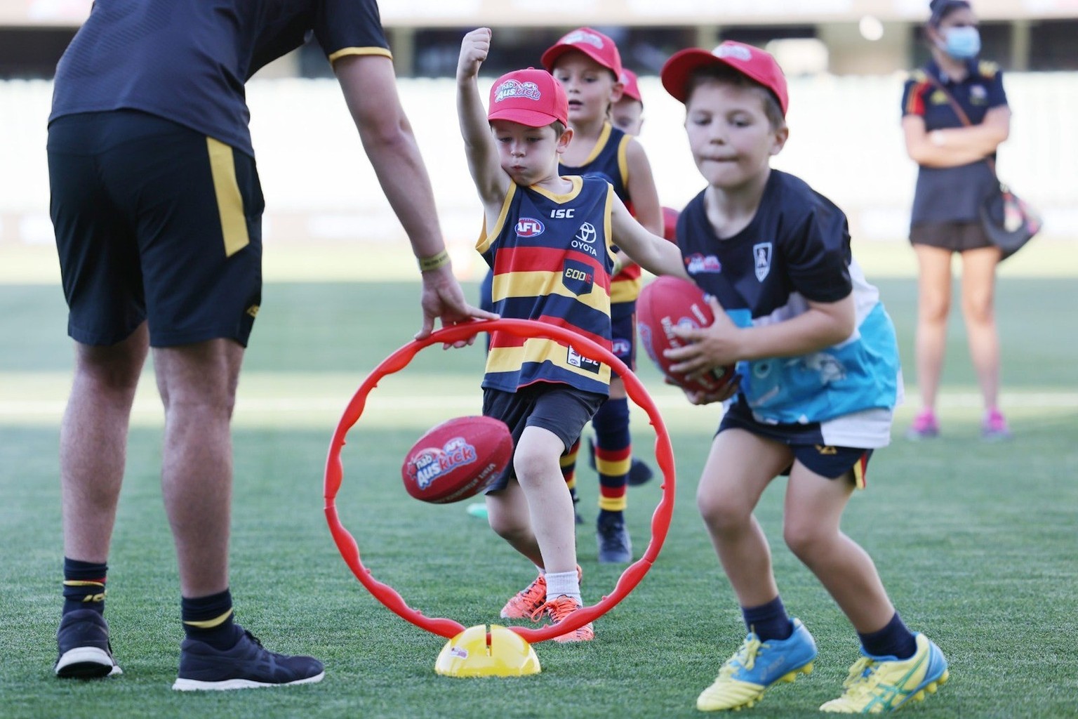 Young children in AFL guernseys kick footballs during an AusKick clinic on a stadium ground