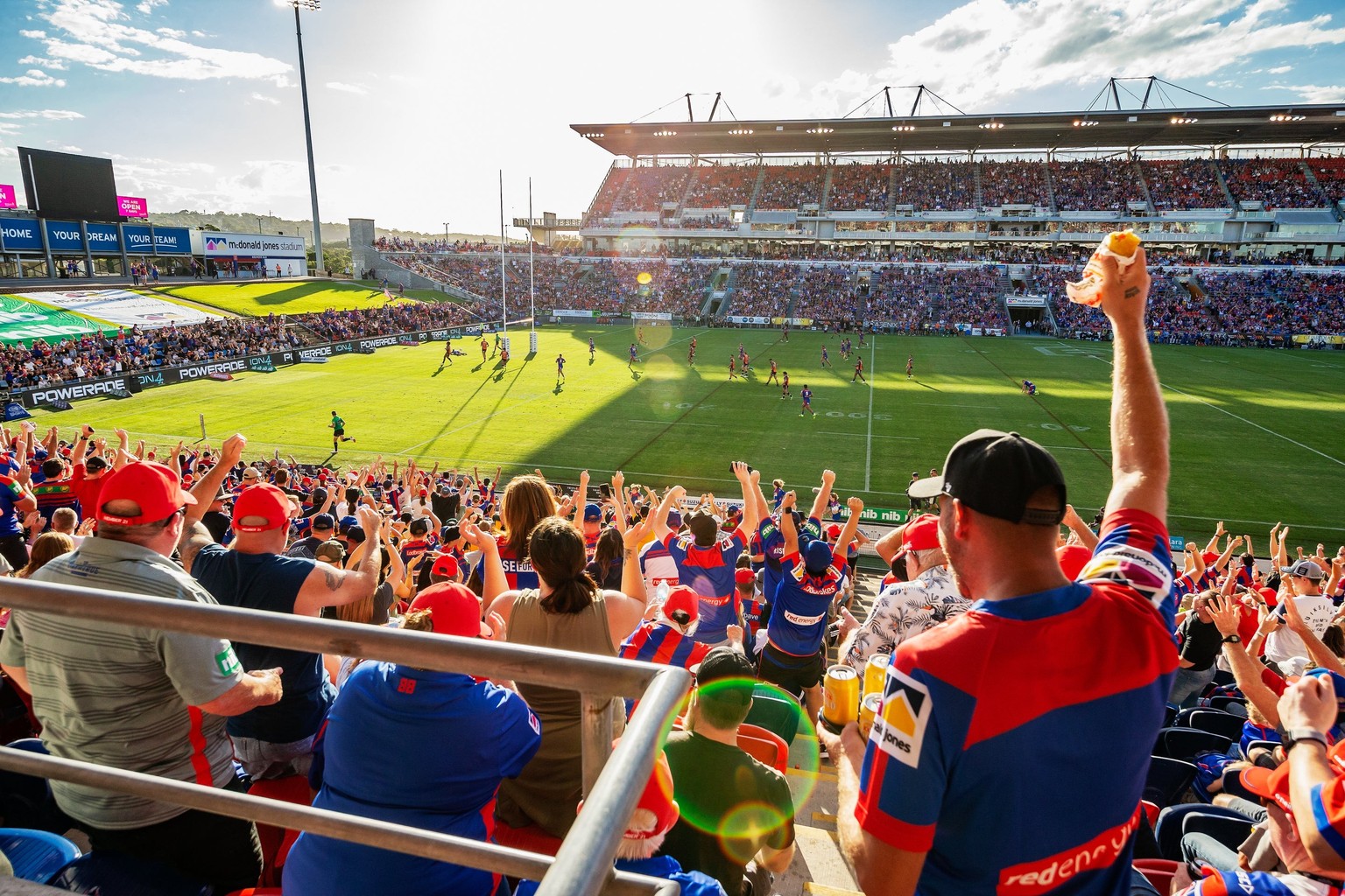 Newcastle supporters in red and blue standing and cheering in the foreground at McDonald Jones Stadium, the match going on behind them