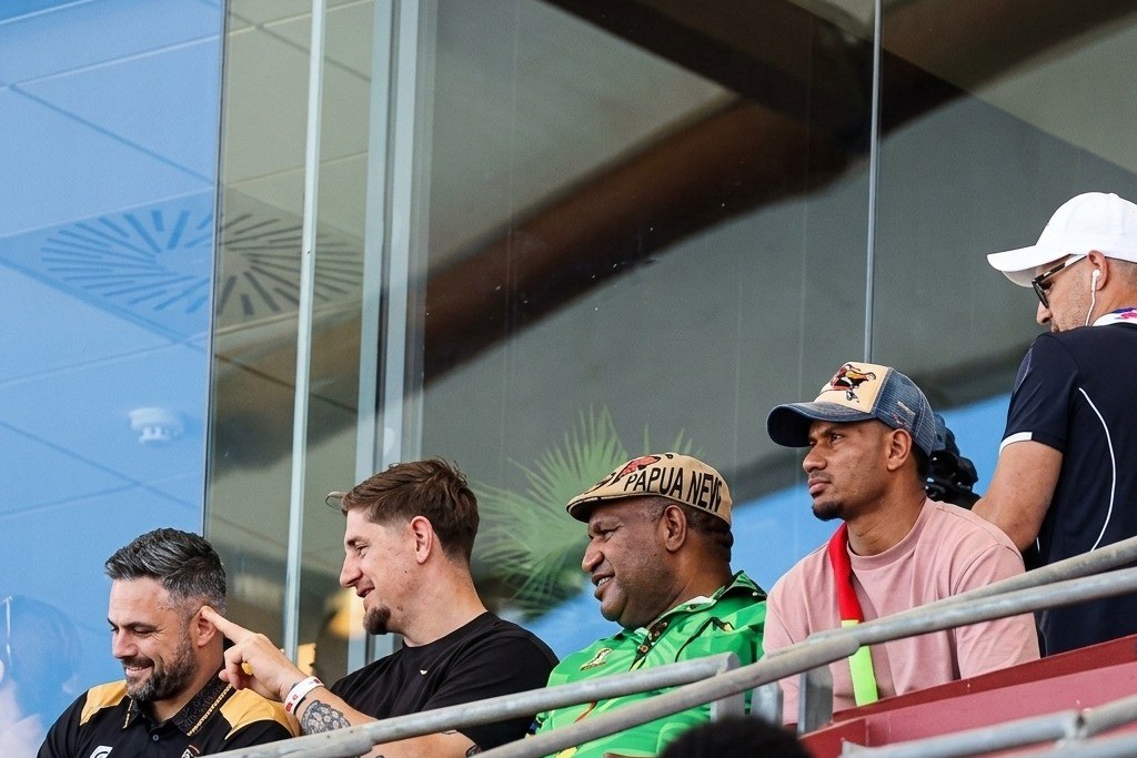 Papua New Guinea prime minister James Marape, in a Papua New Guinea cap, watches a rugby league match from the stands alongside officials and players