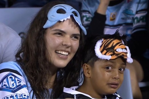 A young Wests Tigers fan in a tiger mask cries in the crowd beside a smiling Cronulla Sharks supporter