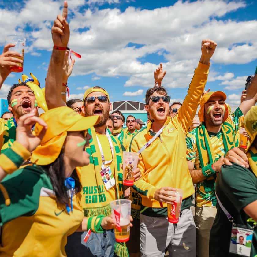A crowd of Australian sports fans in green and gold, cheering with drinks raised